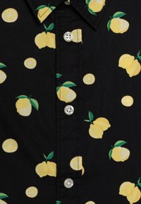 Chemise noire avec un motif citron incluant des citrons entiers et tranchés en jaune ainsi que des feuilles vertes, avec des boutons blancs sur le devant.