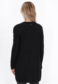 Zwarte gebreide cardigan met een lange, relaxte pasvorm, geribbelde manchetten en een subtiele textuur. Beschikt over een klein logo-label aan de achterkant van de halslijn.