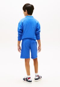 Αγόρι που στέκεται με την πλάτη, φορώντας μπλε φούτερ και αντίστοιχα σορτς, αθλητικά παπούτσια και λευκές κάλτσες, σε απλό λευκό φόντο.