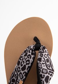 Zwarte en grijs gemustreerde stof thongband met een knoop bovenop een bruine, gladde, gewatteerde zool, ontworpen voor comfort en stabiliteit.