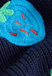 Blaue gehäkelte Erdbeere mit roten Samen und grünen Blättern auf dunkelblauem Strickstoff.