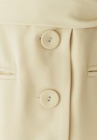 Bottoni di un cappotto in tessuto beige, con bottoni rivestiti in tessuto abbinato e tasche a fessura su entrambi i lati.