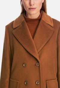 Brown double-breasted coat in textured fabric, featuring a wide collar, two front pockets, and dark brown buttons.