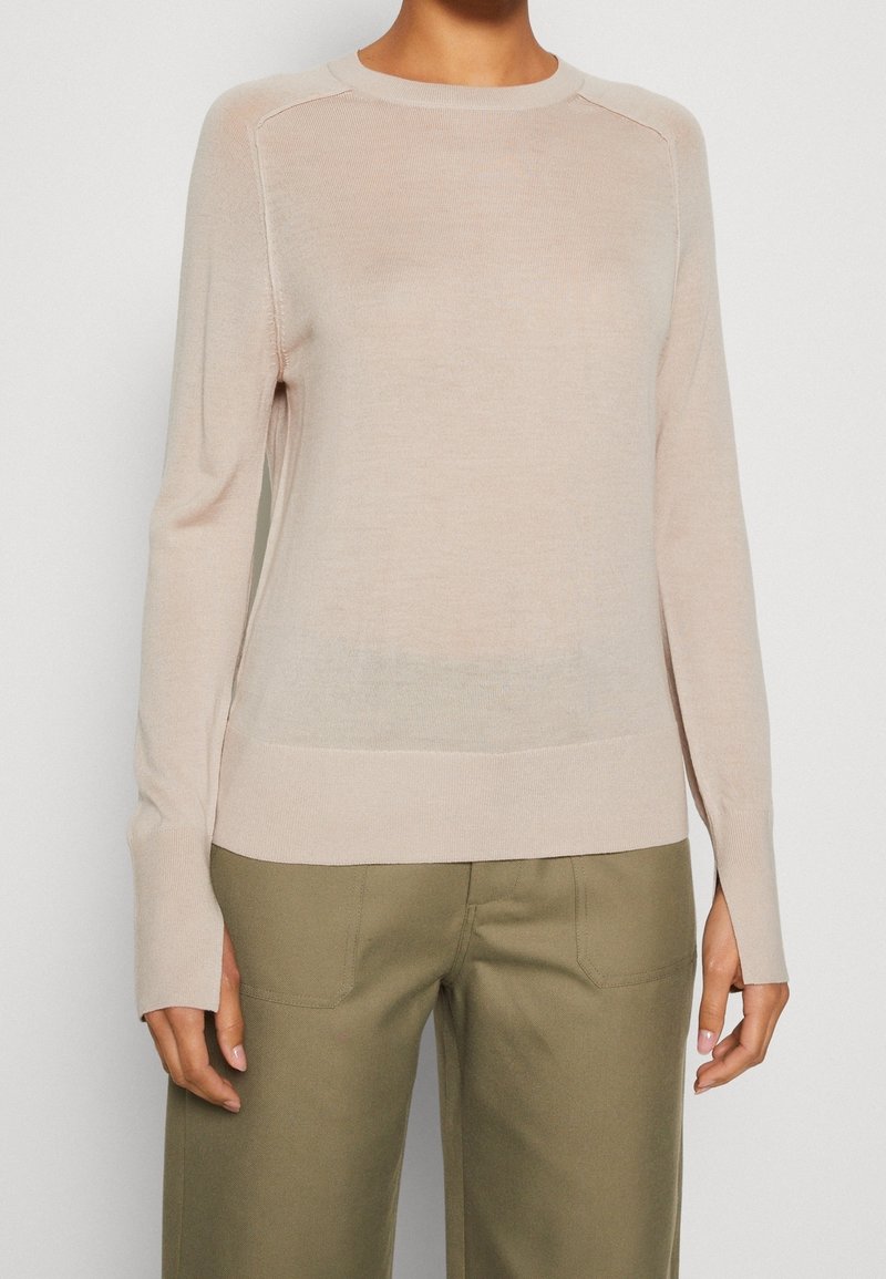 Helle beige Strickjacke mit langen Ärmeln, geripptem Halsausschnitt und Saum, mit einer subtilen durchsichtigen Textur und engen Bündchen. Kombiniert mit olivgrünen Hosen.