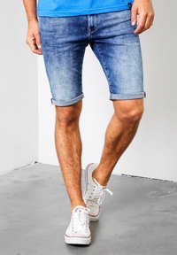 Shorts de mezclilla azul claro con dobladillos enrollados, con un ajuste clásico y un leve desgaste. Llevados con zapatillas blancas.