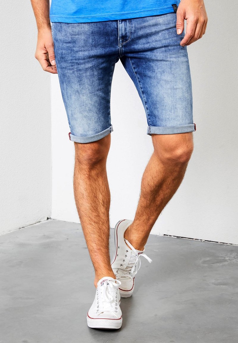 Shorts de mezclilla azul claro con dobladillos enrollados, con un ajuste clásico y un leve desgaste. Llevados con zapatillas blancas.