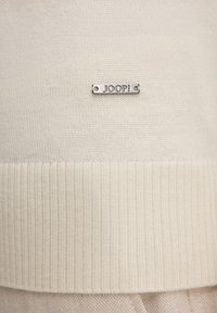 Maglione a maglia color crema con orlo a coste e una targhetta logo argentata con la scritta "JOOP!" in rilievo. Tessuto morbido, design semplice.