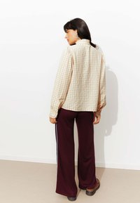 Femme aux cheveux bruns portant un blouse à volants beige à carreaux, un pantalon large bordeaux avec des rayures blanches et des chaussures à imprimé léopard, se tenant de côté.