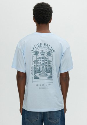 Hombre con camiseta azul claro con gráfico detallado de edificio y palmeras y texto "Azure Palms Resort & Spa" en la espalda.