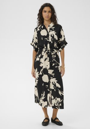 Woman with curly hair wearing a black and cream floral button-up midi dress and black flat shoes, standing against a plain background.
