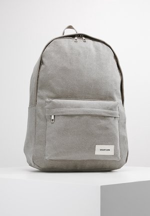 Sac à dos en tissu gris à forme arrondie, avec une poche avant zippée et une petite étiquette logo blanche. Texture lisse et design minimaliste.