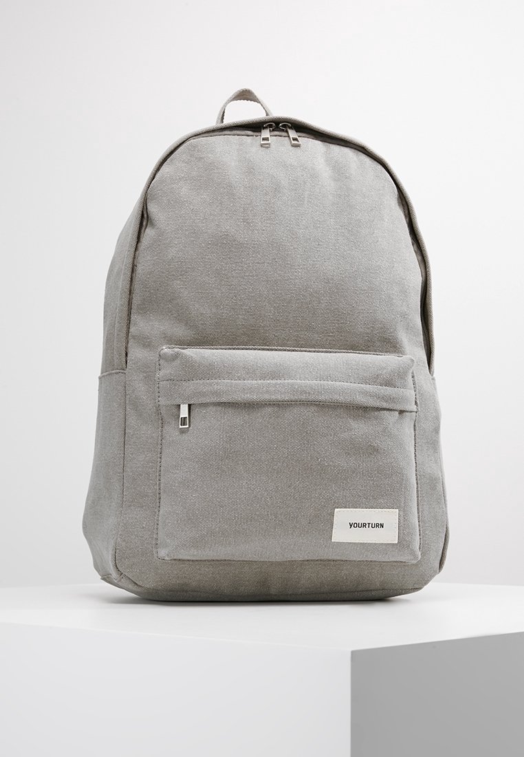 Sac à dos en tissu gris à forme arrondie, avec une poche avant zippée et une petite étiquette logo blanche. Texture lisse et design minimaliste.
