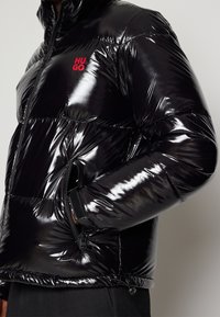 Osoba ubrana w błyszczącą czarną kurtkę puchową z czerwonym logo "HUGO" na klatce piersiowej oraz kieszeniami na zamek błyskawiczny z boku, z rękami w kieszeniach.