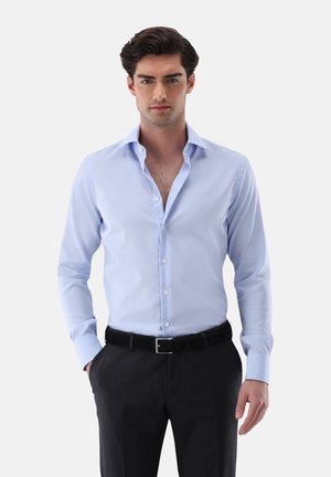 Camicia da uomo in cotone azzurro chiaro con colletto strutturato, chiusura frontale con bottoni e maniche lunghe, abbinata a pantaloni scuri e cintura nera.
