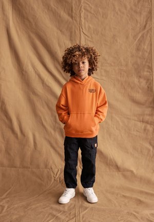 Bambino con capelli ricci indossa felpa arancione con cappuccio, pantaloni cargo neri e sneakers bianche, in piedi davanti a uno sfondo beige in tessuto.