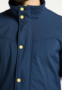 Mörkblå jacka med hög krage, prydd med gula knappar, slät textur och sömmar längs bröstet och axlarna.