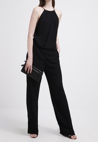 Combinaison noire dos nu à silhouette à jambes larges, en tissu doux avec des détails plissés ; accompagnée d'une pochette noire texturée.