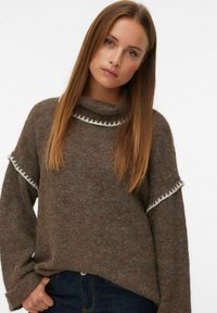 Pull marron en laine avec un col montant, doté d'une bordure blanche à volants sur les manches. Coupe légèrement ample et tissu texturé.
