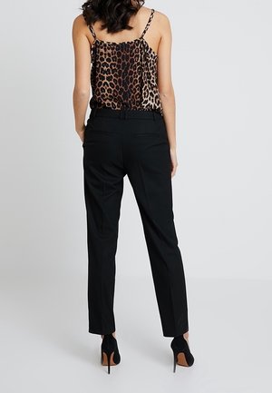 Femme portant un pantalon noir ajusté, des talons hauts noirs et un haut sans manches imprimé léopard, debout, les mains dans les poches, dos tourné.