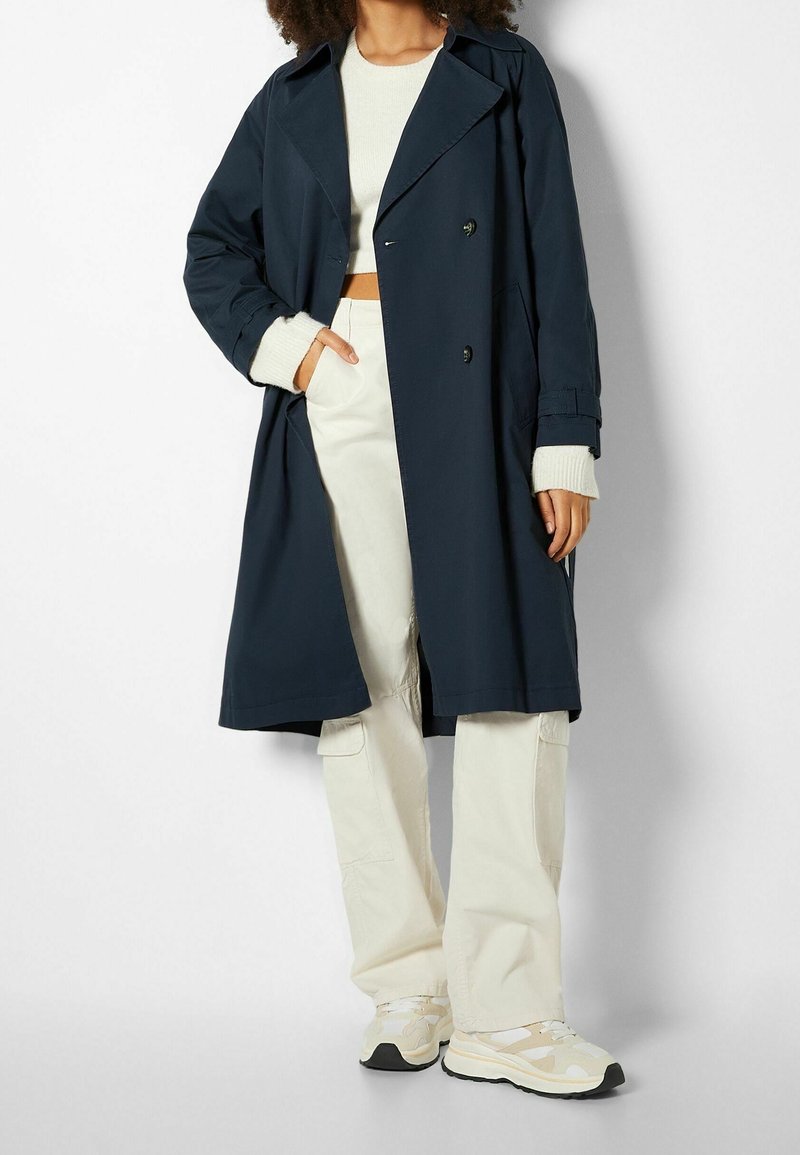 Trench-coat bleu marine à double boutonnage, larges revers et boutons décoratifs. Porté sur un pull léger et un pantalon cargo crème.