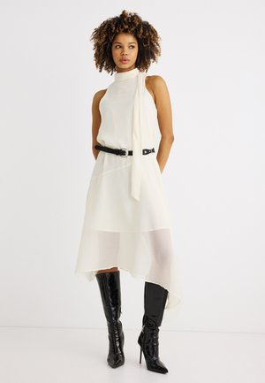 Frau mit lockigem Haar, die ein weißes ärmelloses Kleid mit hohem Kragen und asymmetrischem Saum, schwarzen Gürtel und schwarze kniehohe Stiefel mit Absatz trägt.
