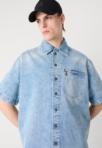 Camisa de mezclilla de manga corta color azul claro con cierre de botones en la parte delantera, bolsillo en el pecho y botones oscuros. Presenta un ligero desvanecimiento y costuras de contraste.