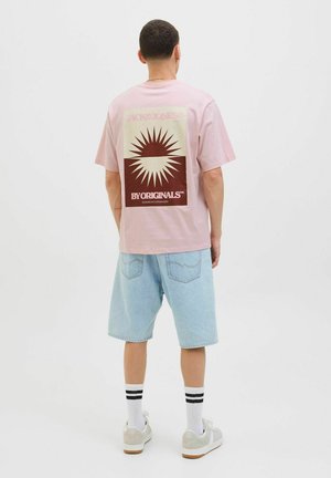 Roze T-shirt met korte mouwen en een grafisch ontwerp met een bruine zonnestraalmotief op de rug, gecombineerd met lichtblauwe denim shorts.
