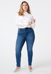 Chemise blanche à boutons avec de petits accents décoratifs, associée à un jean en denim bleu ajusté et des chaussures à talons argentées brillantes.