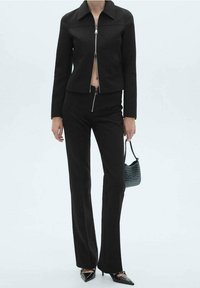 Veste noire ajustée avec une fermeture éclair devant et un col, assortie à un pantalon noir taille haute. Comprend un petit sac à main vert texturé et des chaussures pointues.