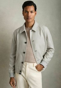Chaqueta de tejido houndstooth gris con cuello tipo polo, cierre de botones y puños acanalados. Usada sobre una camisa de color beige claro y pantalones crema.