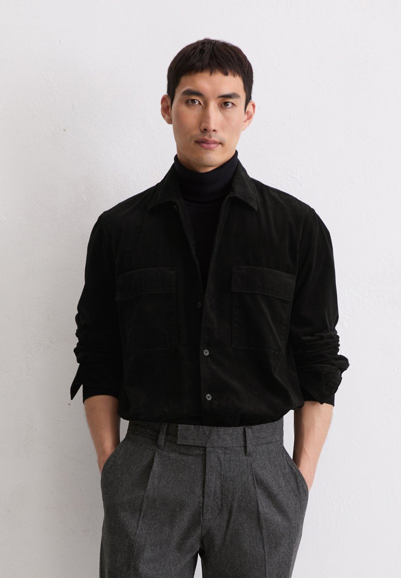 Uomo che indossa una camicia di velluto a coste nera sopra un dolcevita nero, con pantaloni grigi a pieghe, in piedi con le mani in tasca contro un muro bianco.