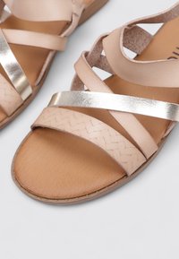 Sandalias de cuero beige con diseño de tiras cruzadas, acentos metálicos, detalles trenzados y suelas planas. Textura suave con un aspecto casual.