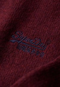 Dunkelroter gestrickter Stoff mit navyblauer gestickter SuperDry-Logo, das aus Schrifttext und einem stilisierten Flügeldesign besteht.