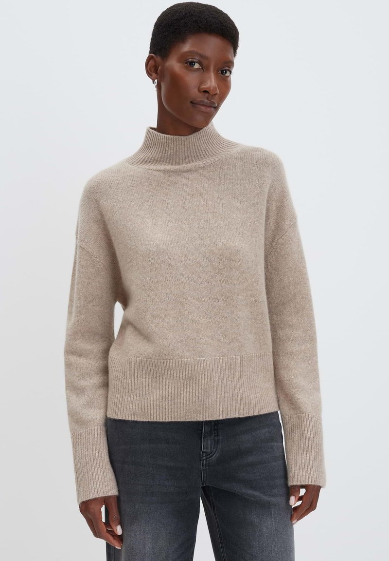 Beiger Strickpullover mit Rollkragen, geripptem Saum und Ärmeln, lässiger Schnitt und weicher Textur. Getragen mit dunkelgrauen Jeans.