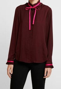 Blusa bordeaux a maniche lunghe con scollo plissettato, fiocco in raso rosa e polsini strizzati rosa. Tessuto morbido con chiusura frontale a bottoni.