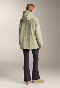 Osoba v svetlo zelenej prešívanej bunde s kapucňou, tmavých rozšírených nohaviciach a béžových platformových topánkach, stojaca chrbtom na neutrálnom pozadí.