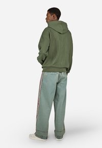 Zelená rebrovaná mikina s kapucňou na šnúrku, v kombinácii so svetlomodr�ými širokými džínsami s bočnými pruhmi a bielymi teniskami.