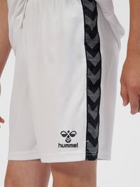 Hvide atletikshorts med sort chevron-mønstret stribe på siden og sort "hummel" logo nær kanten, båret af en person.