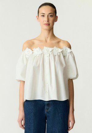 Kvinde iført en hvid bluse med bar skuldre og blomsterapplikationer langs udskæringen og blå denimjeans, stående mod en ensfarvet baggrund.