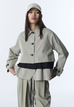 Mujer joven con chaqueta beige en capas, pantalones anchos a juego y una gorra de béisbol beige, de pie con las manos en la cadera sobre un fondo blanco.