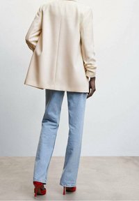 Béžový blejzr s volným střihem, dlouhými rukávy a rozparkem vzadu, kombinovaný se světle modrými zvonovými džíny a červenými vysokými podpatky.