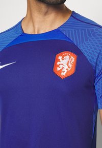 Camiseta atlética morada con hombros azules estampados, que presenta un logo en forma de escudo naranja con un león y texto. Hecha de material transpirable.