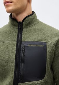 Homme portant une veste polaire verte avec une poche poitrine zippée noire étiquetée "PeakPerformance" sur un fond uni.