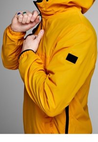 Žlutá voděodolná bunda s kapucí, s černými detaily a elastickými manžetami. Tkanina vypadá hladká a lehká.