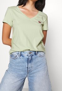 Jasnozielony T-shirt z dekoltem w serek wykonany z miękkiego materiału. Posiada krótkie rękawy oraz małe czerwone logo Levi's na piersi, zestawiony z wyblakłymi niebieskimi dżinsami.