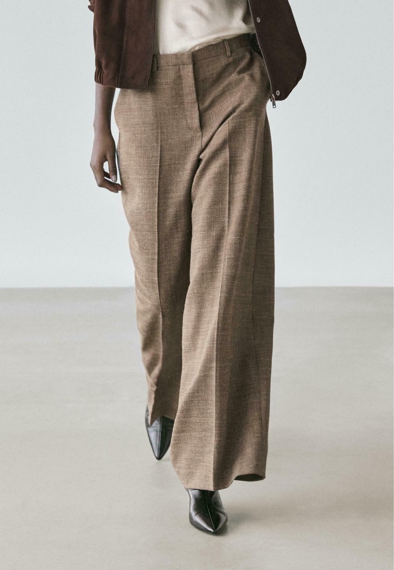 Personne portant un pantalon marron à jambes larges, des chaussures noires à bout pointu, un haut blanc et une veste sombre, marchant sur un sol clair.