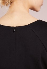 Mustaha dress musta, jossa on leveä kaula-aukko, takana vetskari ja sileä, tekstiilinen kangas. Suunnittelussa on hienovaraista sauman yksityiskohtaisuutta hartioilla.