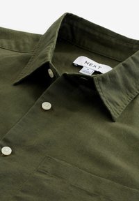 Chemise boutonnée olive avec col, boutons blancs et une étiquette en tissu indiquant la taille M à l’intérieur du col.
