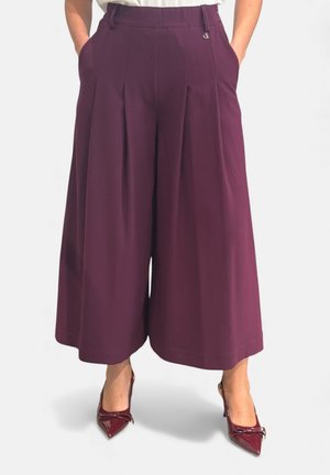 Donna che indossa culottes prugna a vita alta e gamba larga con pieghe frontali e décolleté abbinate a punta con fiocchi. Mani nelle tasche.