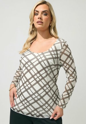 Femme aux longs cheveux blonds portant un haut blanc à manches longues avec un motif à carreaux marron, des boucles d'oreilles créoles dorées et un pantalon noir.